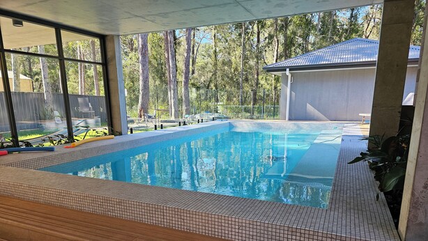 A swimming pool with tiled edges, wooden deck, and a view of trees and a building in the background. - Sanctuary on the Creek (Sanctuary Point)