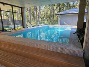 A swimming pool with tiled edges, wooden deck, and a view of trees and a building in the background. - Sanctuary on the Creek (Sanctuary Point)