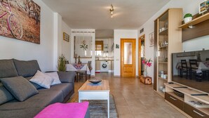 Un salon avec un canapé, une table basse et un meuble pour la télévision. On y trouve également un coin repas avec une table et des chaises, et une cuisine à l’arrière-plan.