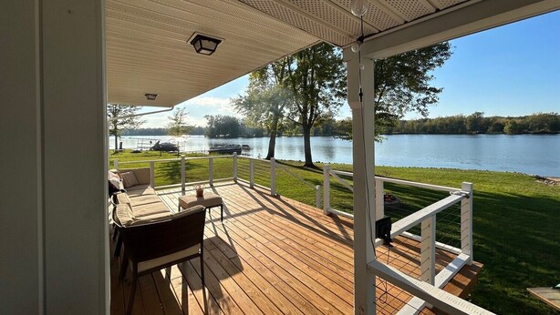 A covered deck with a view of a lake, a table with chairs, and a railing.