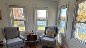 A room with hardwood flooring, two grey armchairs, a small wooden table, and large windows offering a view of a lake and trees.