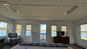 A bedroom with a large bed, a chair, a dresser, a television, and multiple windows offering a view of a lake.