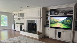 A spacious living room with a large flat-screen TV, built-in bookshelves, a fireplace, and a view of the outdoors.
