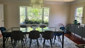 A dining area with a glass table, four chairs, a modern chandelier, and a view of greenery through large windows.