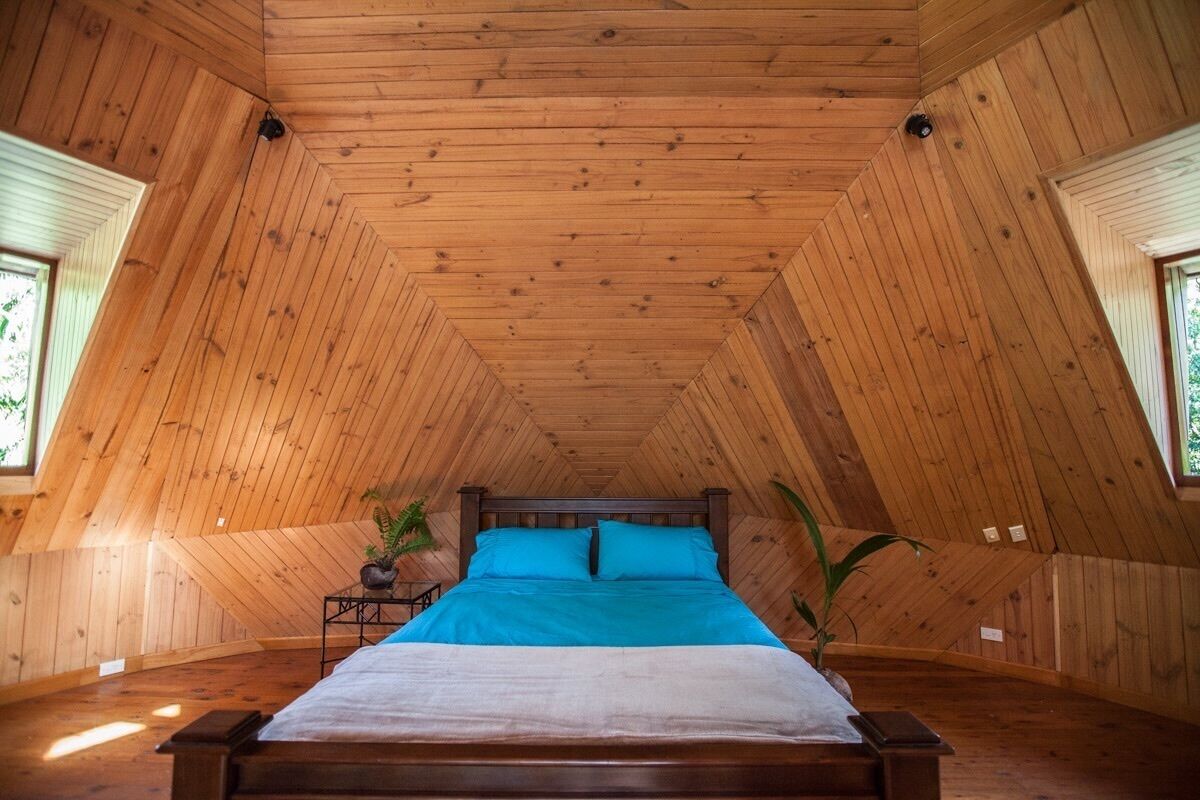 Quarto com teto inclinado de madeira, cama com roupa de cama azul e duas janelas em ambos os lados.