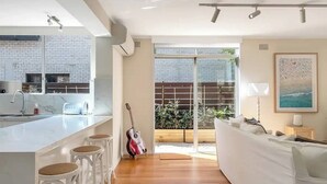 A modern kitchen with a white island and bar stools, a dining area with a white sofa, and a view of a brick wall and greenery outside. - Luxe-Coastal Balmoral Townhouse
 (Mosman)
