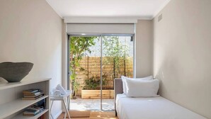 A modern bedroom with a bed, a white shelf with books, a small table with a towel, and a sliding glass door leading to a bamboo garden. - Luxe-Coastal Balmoral Townhouse
 (Mosman)