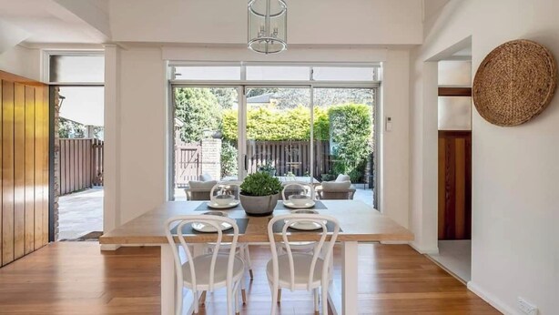 A dining area with a wooden table, white chairs, and a hanging light fixture. - Luxe-Coastal Balmoral Townhouse
 (Mosman)