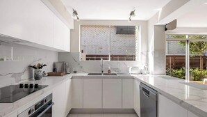 A modern kitchen with white cabinetry, a built-in oven, a dishwasher, and a window with a view of greenery. - Luxe-Coastal Balmoral Townhouse
 (Mosman)