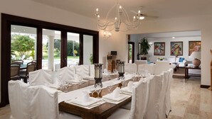 A dining area with a long table set for a meal, large windows offering a view of the outdoors, and a ceiling fan.