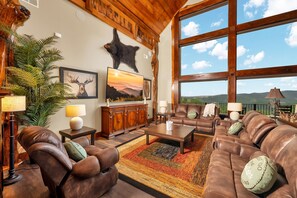 A spacious living room with leather sofas, a large window offering a mountain view, and a bear head mounted on the wall. - Excalibur: Pickleball, Duck Pin Bowling, Indoor Pool, 9 Hole Golf (Sevierville)