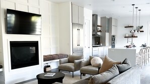 Un salon moderne doté d’un grand téléviseur à écran plat, d’un ventilateur de plafond et d’une cuisine en arrière-plan.