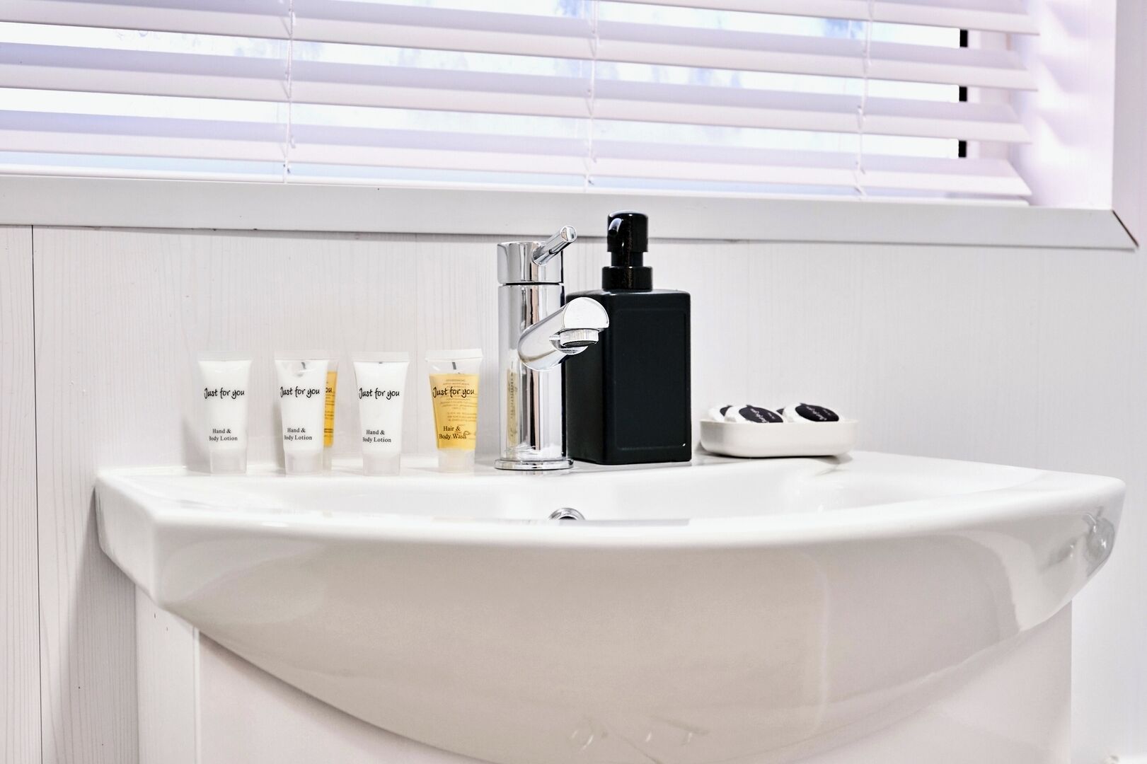 Ett badrum med ett vitt handfat, en svart tvåldispenser och tre tuber lotion med etiketten ”Bara för dig”.