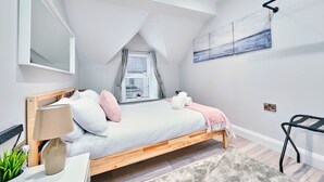 Een moderne slaapkamer met een schuine plafond, een bed met wit beddengoed en roze kussens, een bedlampje, een raam met jaloezieën en een schilderij aan de muur.