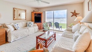 A living room with a large window overlooking the ocean, a ceiling fan, and a wicker coffee table. - Oceanfront Oasis: 2BR Condo w/ Stunning Views (New Smyrna Beach)