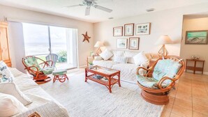 A living room with wicker furniture, a glass coffee table, and a view of the ocean through large windows. - Oceanfront Oasis: 2BR Condo w/ Stunning Views (New Smyrna Beach)