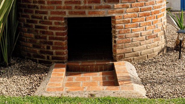 A brick oven structure with a small entrance, surrounded by gravel and grass, with a metal framework and pipes above. - Glamping Cholula (San Pedro Cholula)