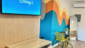 Een moderne kantoorruimte met een houten bureau, een grijze bureaustoel, een kleurrijke muur, een flatscreen televisie waarop het Disney+-logo staat, een gele stoel en een klein tafeltje met een plant.