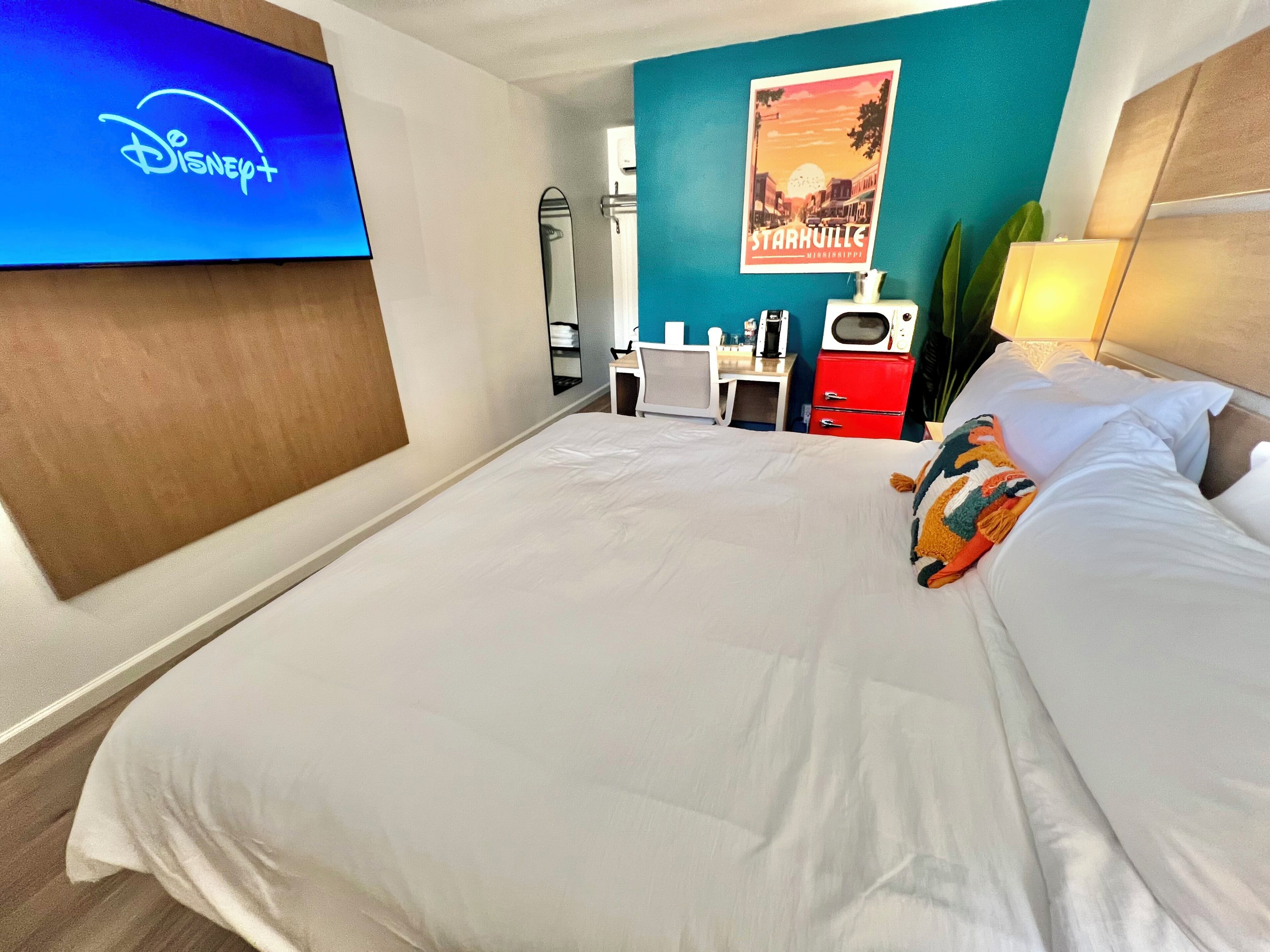 Una habitación de hotel con una cama, un televisor mostrando el logotipo de Disney+, un cartel de Starkville, Mississippi, y un escritorio con una cafetera.