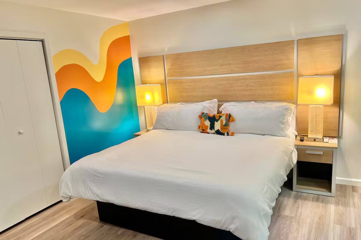 A hotel room with a large bed, two bedside tables with lamps, a colorful wall mural, and a wooden headboard.
