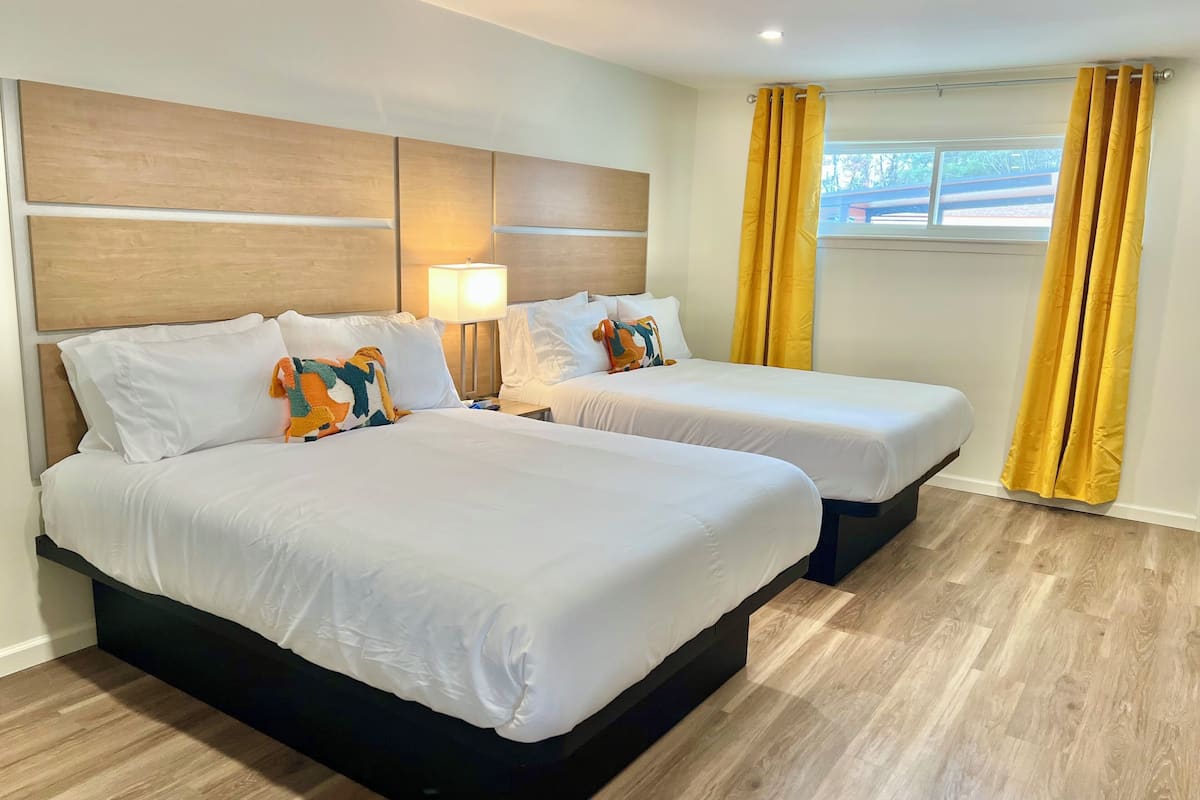 A hotel room with two beds, a nightstand, a lamp, and a window with yellow curtains.