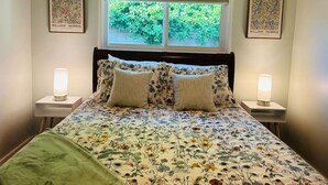 Une chambre à coucher avec une couvre-lit à motifs floraux, deux lampes de chevet, une fenêtre avec des stores et deux œuvres d’art encadrées accrochées aux murs.