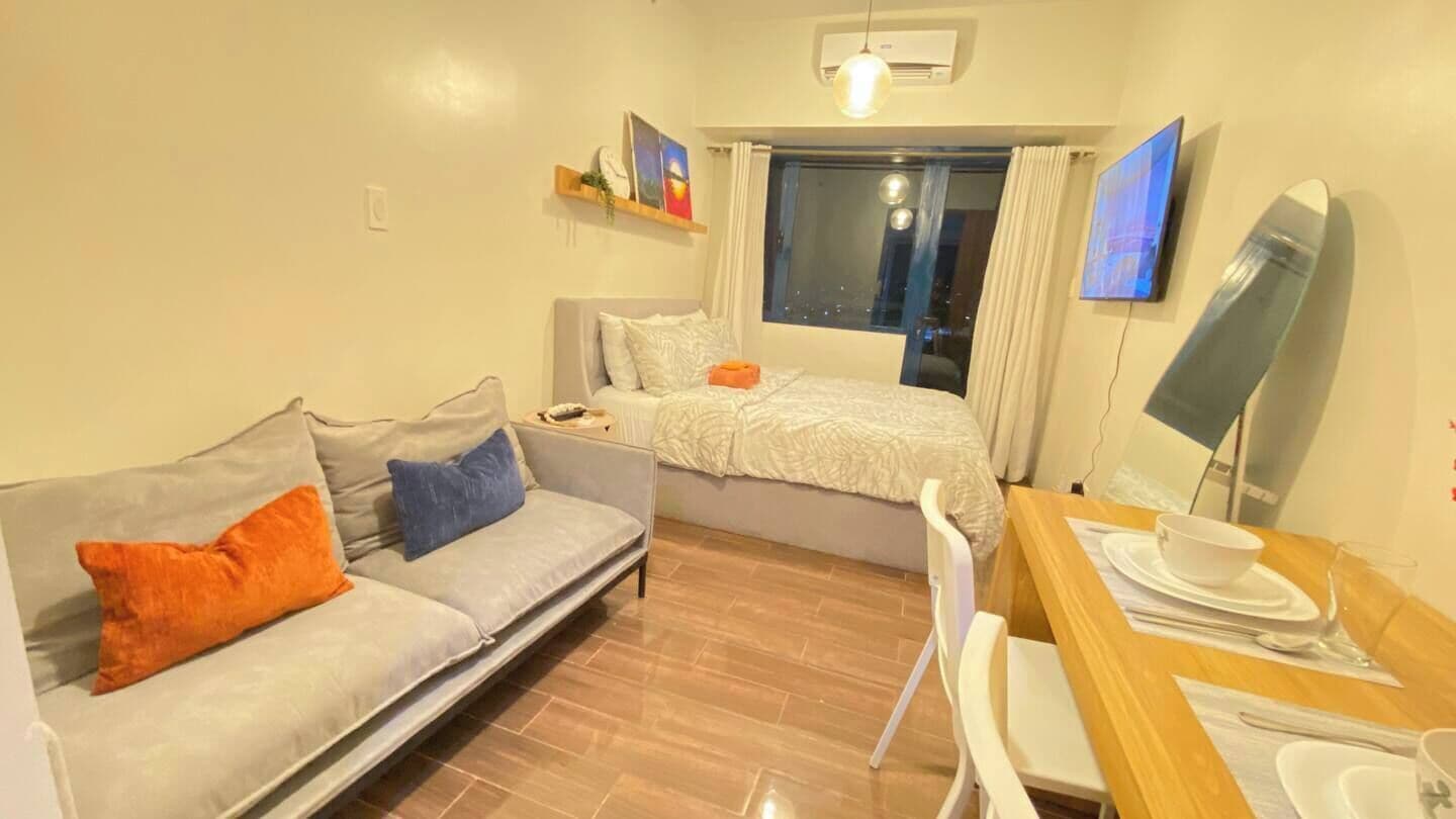A compact hotel room with a sofa, a bed, a dining table with chairs, a TV, and a window with curtains.