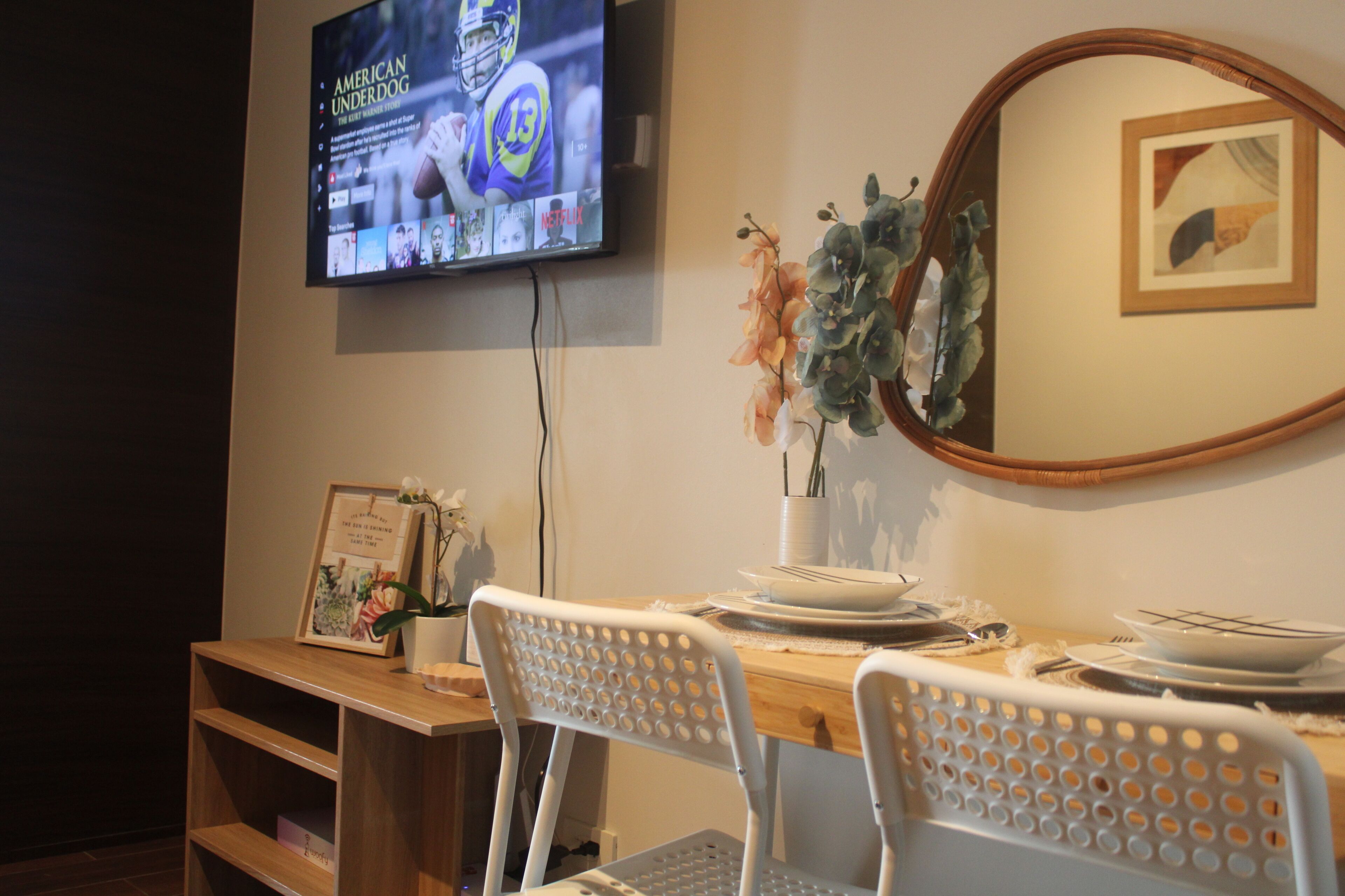 Un comedor con una mesa de madera, sillas blancas, un televisor montado en la pared y un cuadro enmarcado.