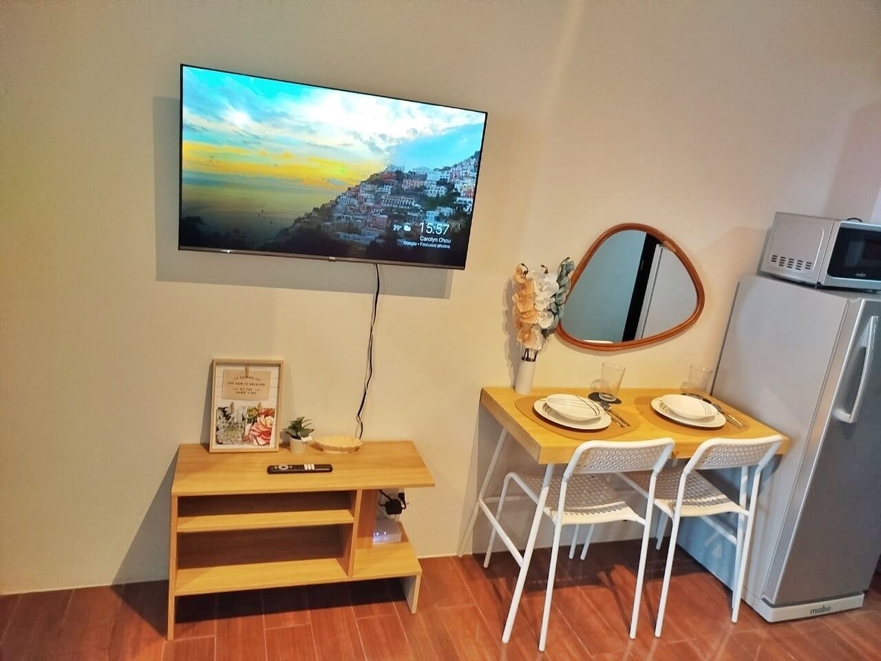 A compact living space with a TV mounted on the wall displaying a scenic view, a small wooden table with chairs, a microwave, and a refrigerator.