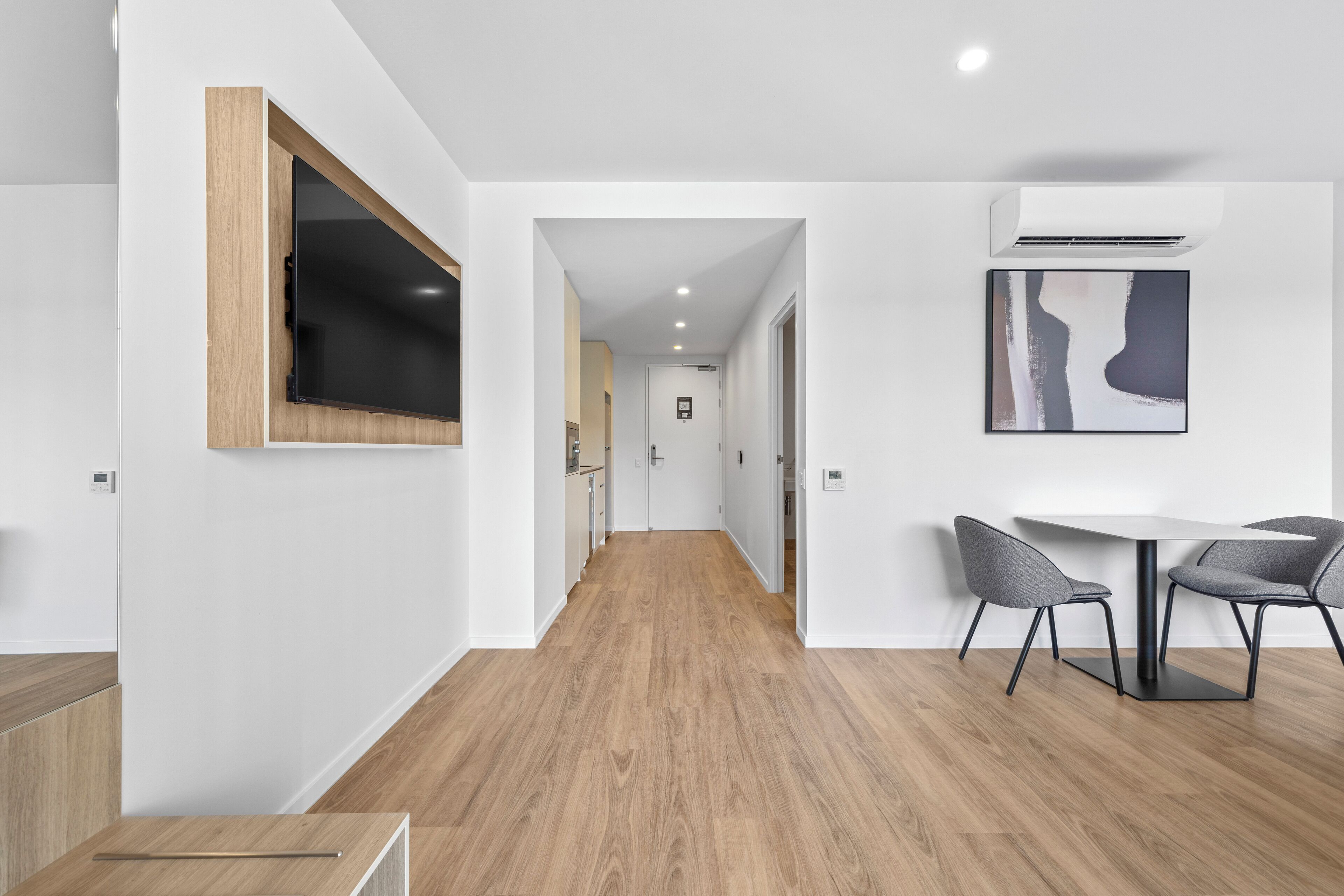 A modern interior with a wooden floor, a dining area with two chairs, a flat-screen TV mounted on a wooden panel, and a framed artwork on the wall.