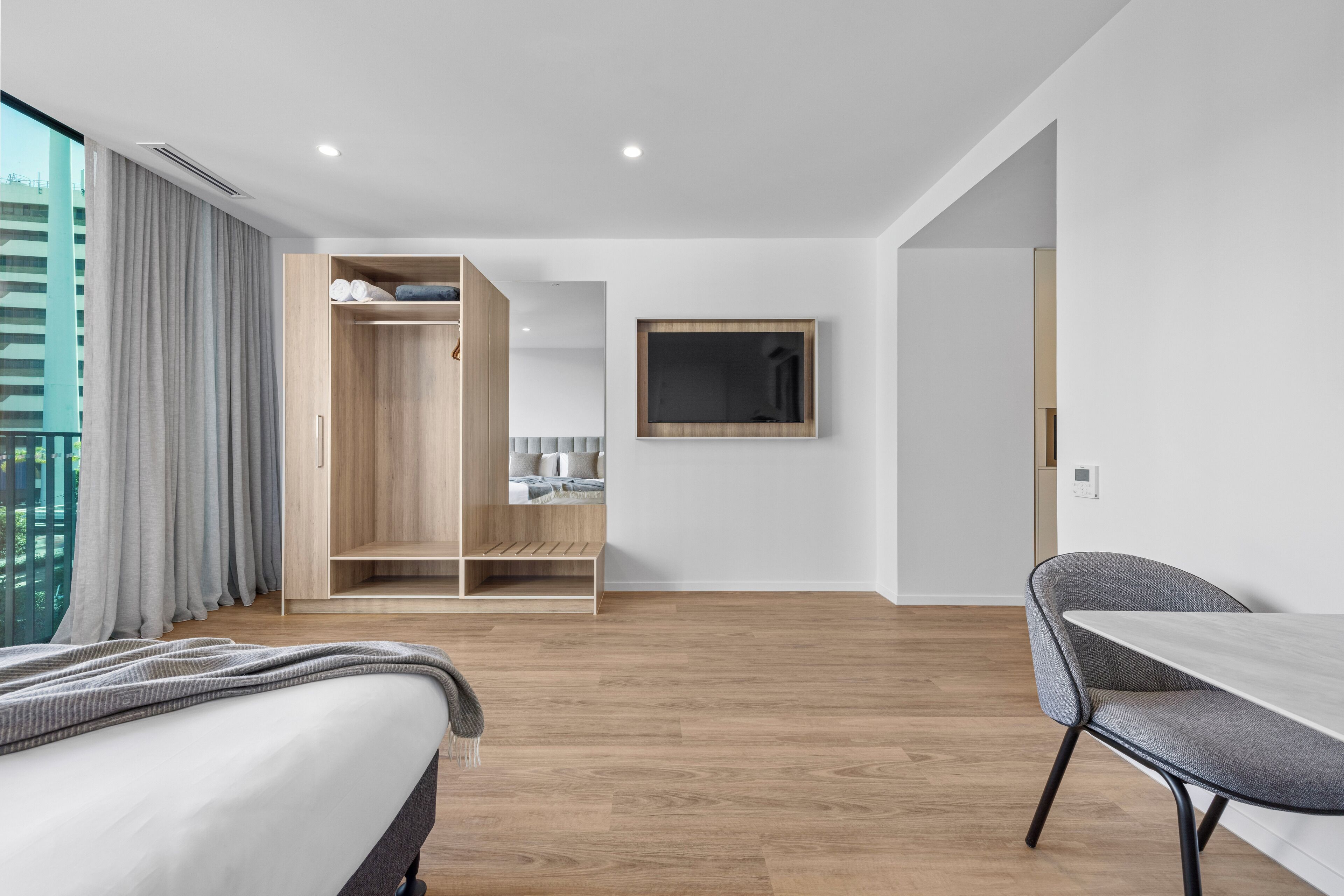 Ein modernes Hotelzimmer mit einem Bett, einem grauen Sessel, einem Holzschrank und einem Fernseher an der Wand.
