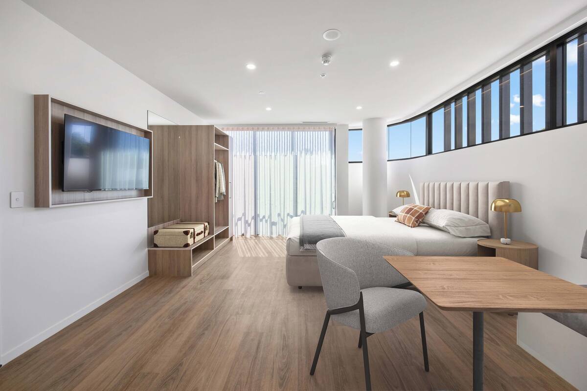 A modern hotel room with a bed, bedside table, lamp, TV, and a seating area with a table and chairs.
