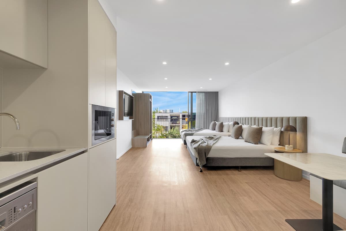A modern hotel room with a kitchenette, a bed with a headboard, a bedside table, and a view of the city.