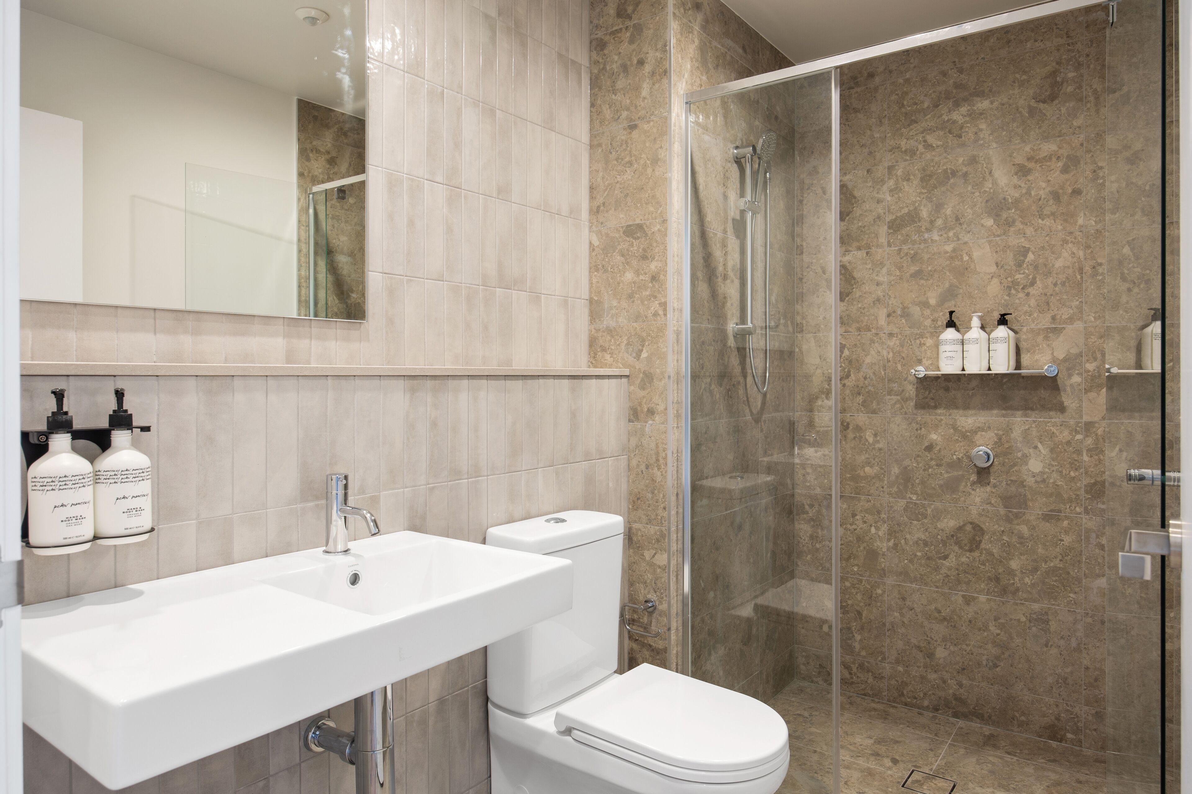 Et moderne badeværelse med en hvid vask, toilet og brusebad. Der er to shampoo flasker på væggen.