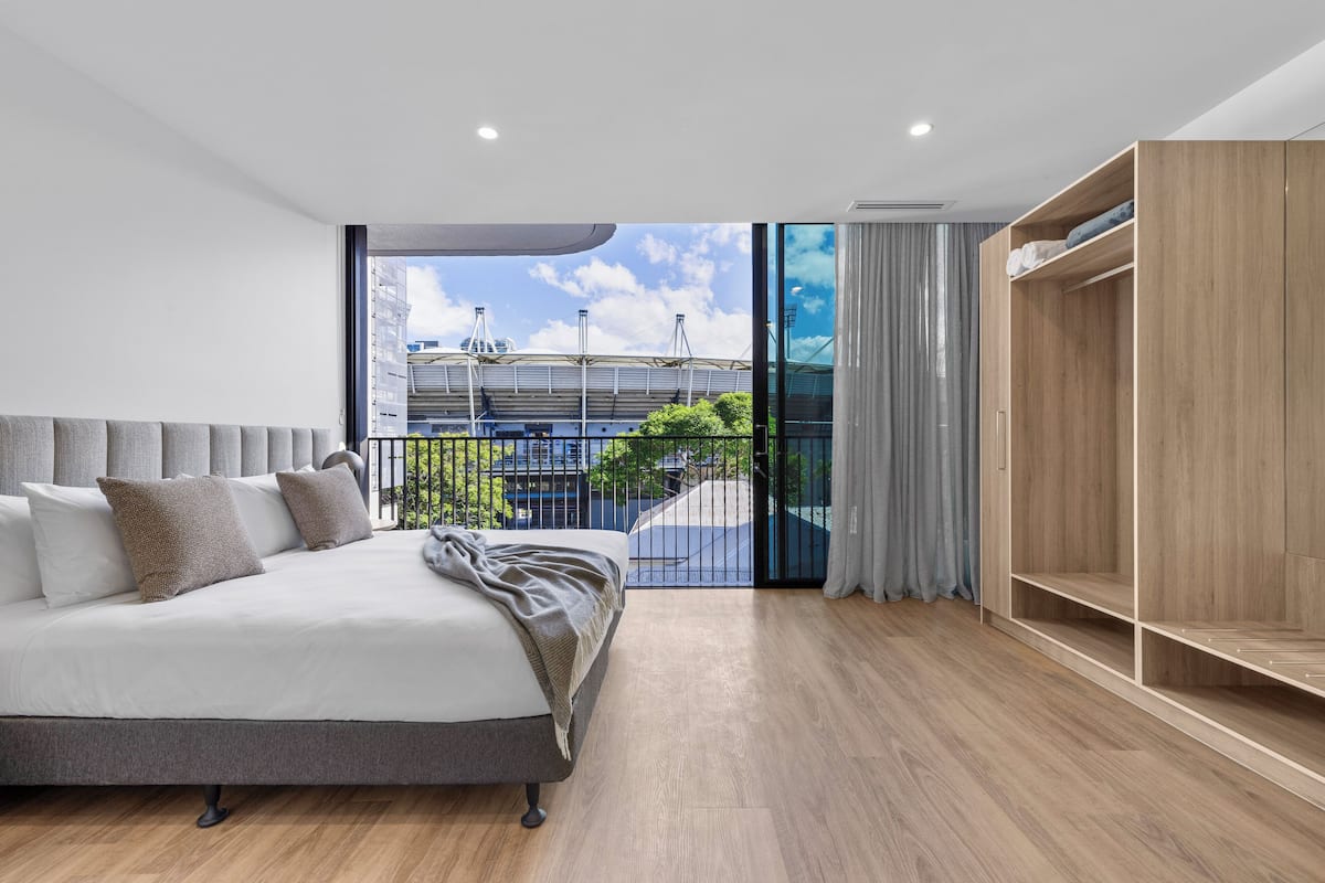 A modern bedroom with a large bed, a wooden wardrobe, and a view of a stadium through large windows.