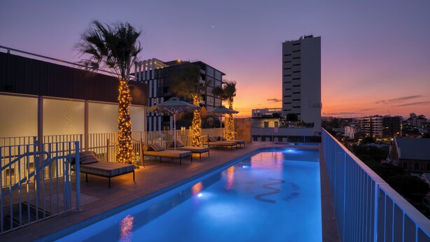A rooftop pool area with lounge chairs and palm trees, surrounded by buildings and a clear sky. - Rambla @ Story House (Kangaroo Point)