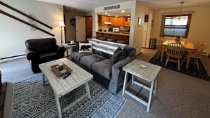 Een woonkamer met een grijze bank, een zwarte lederen fauteuil, een houten salontafel en een klein bijzettafeltje. Er is een eethoek met een houten tafel en stoelen. De keuken is zichtbaar op de achtergrond, met houten kastjes en een raam.
