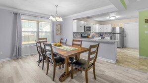 A dining area with a wooden table and chairs, a chandelier, and a view of the kitchen with modern appliances. - Lovely Beach House -Ocean Duplex (Norfolk)