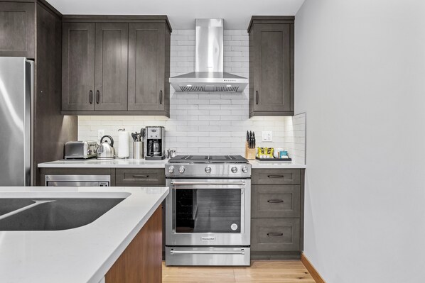A modern kitchen with dark wooden cabinets, a stainless steel range hood, a built-in oven, and a white countertop.