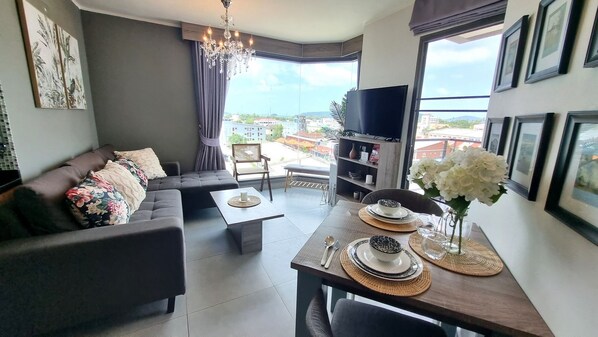 A modern living room with a sofa, dining table, and TV. - Sea View 35 sqm Condo, 6th Floor, WiFi 200 mbps, Pool, Next To Shopping Mall (Chalong)