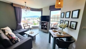 En moderne stue med sofa, sofabord og spisebord. Der er en bogreol, et fjernsyn og en balkon med udsigt over byen.