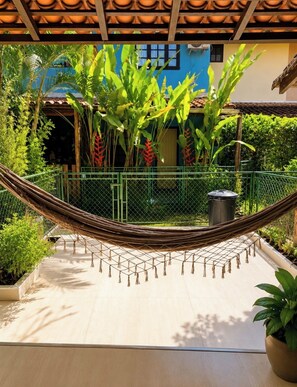 A hammock in a garden with a tiled roof, a trash bin, and various plants.