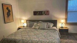 Une chambre à coucher avec une couvre-lit à motifs, deux lampes de chevet, une affiche encadrée au mur et une fenêtre avec des stores.