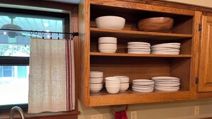 A kitchen with a wooden cabinet containing white and striped bowls and plates, a stainless steel faucet, and a window with a curtain. - Cozy 3-bedroom house in Meridian (Meridian)