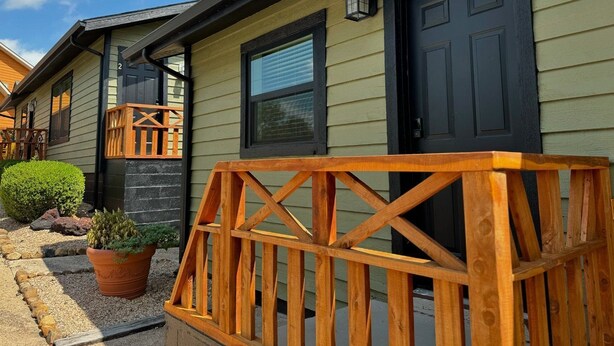 A two-story house with a dark front door, a small porch with a wooden railing, and a potted plant on the ground. - RT 62 Motor Resort '50's Cabin w/Jacuzzi (Eureka Springs)