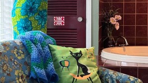 A room with a floral-patterned sofa, a decorative pillow with a cat design, a mannequin in a colorful dress, and a red door with a fire extinguisher sign. - RT 62 Motor Resort '50's Cabin w/Jacuzzi (Eureka Springs)