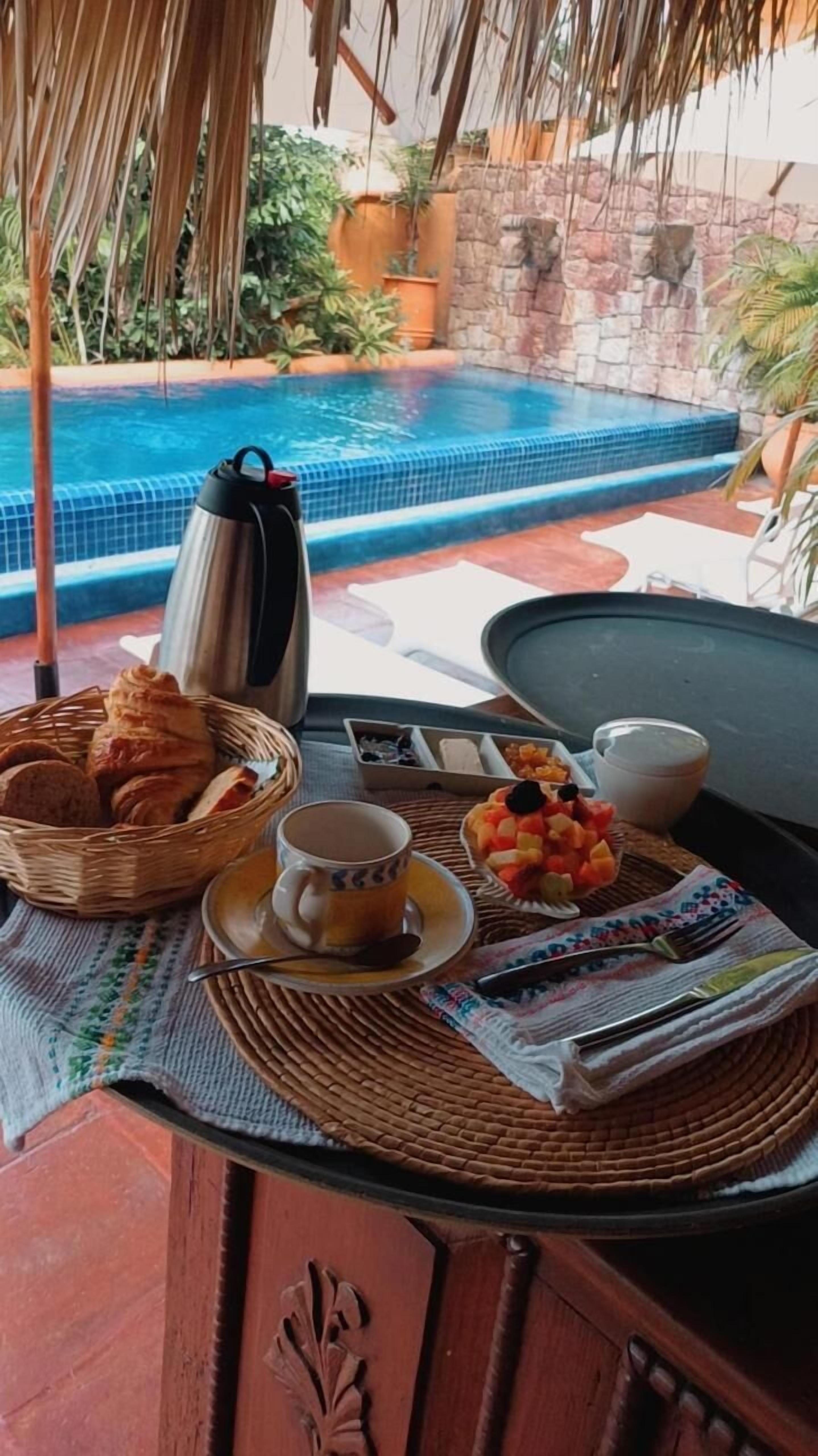 Desayuno con pan, fruta y bebidas sobre una mesa cerca de una piscina.
