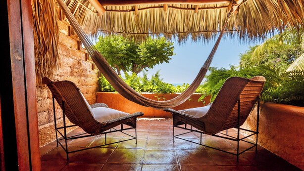 Un hamac tendu entre deux fauteuils en osier sur une terrasse carrelée, avec un mur en briques et un toit de chaume au-dessus.