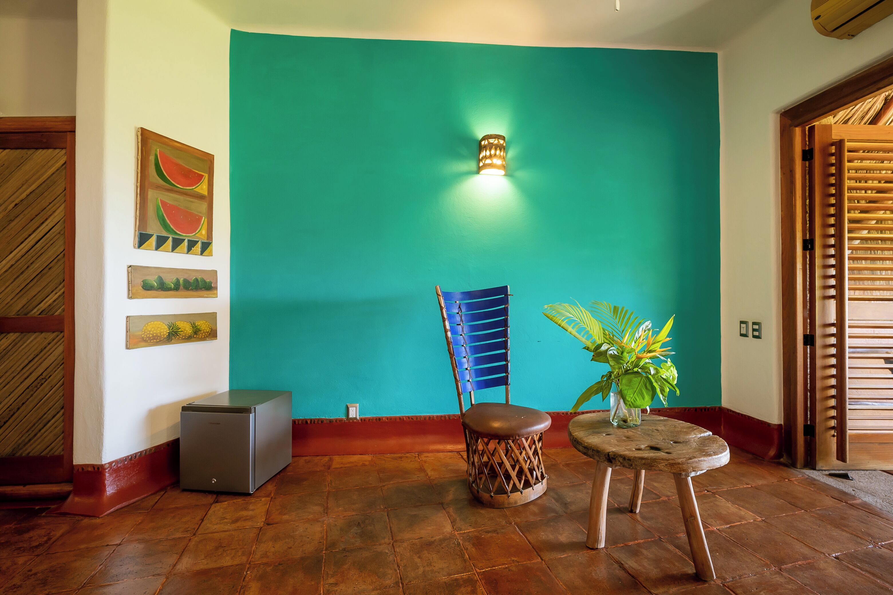 Una habitación con paredes color turquesa, un sillón azul, una mesa pequeña con una planta y un refrigerador.
