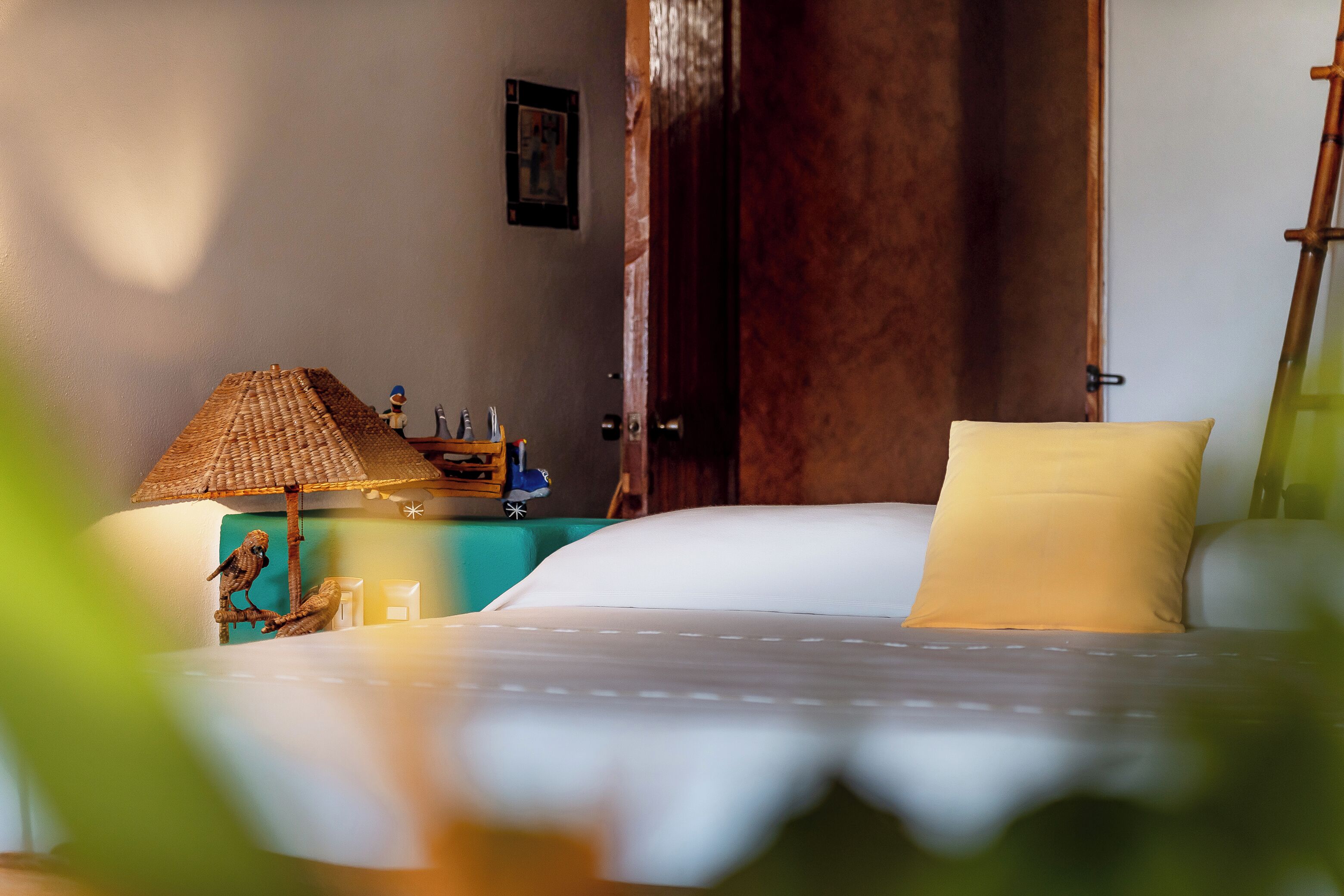 A neatly made bed with a white comforter and a single yellow pillow, a small wooden nightstand with a lamp and a figurine, and a wooden door in the background.